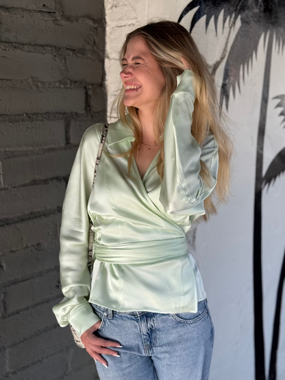 Woman wearing Siena&June light green satin top and jeans standing against a textured wall.